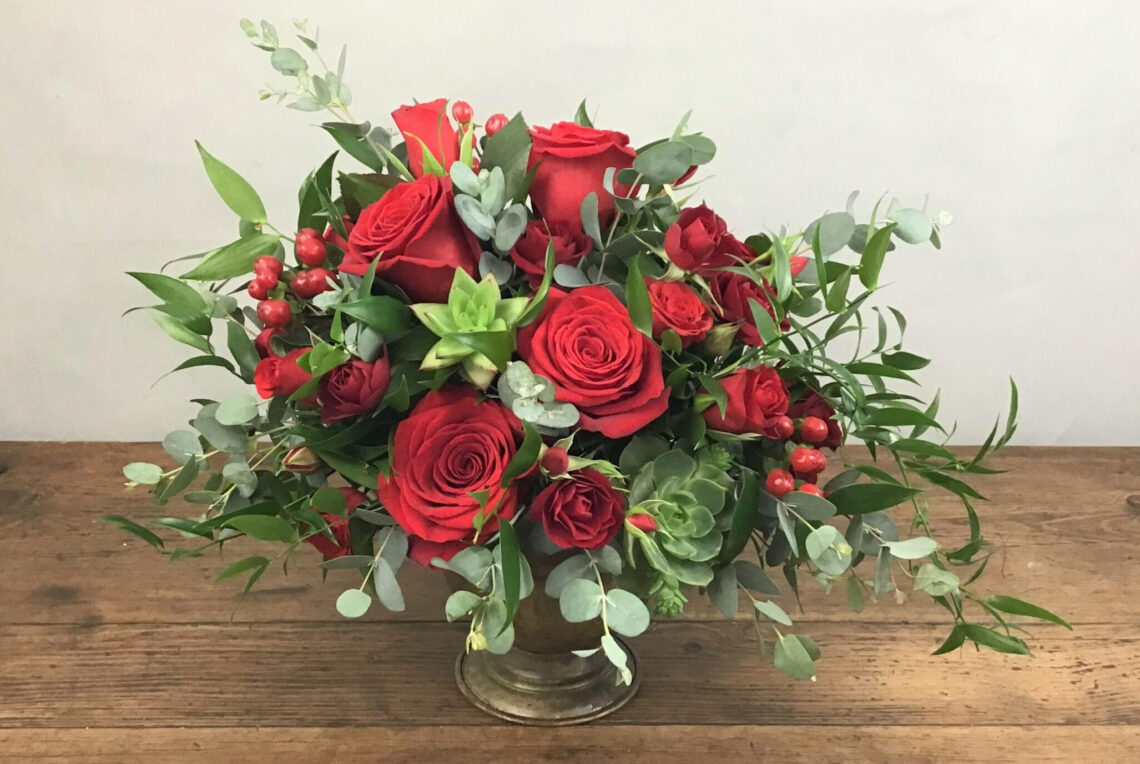 All For Love is red roses with succulents and exotic greens in a compote vase