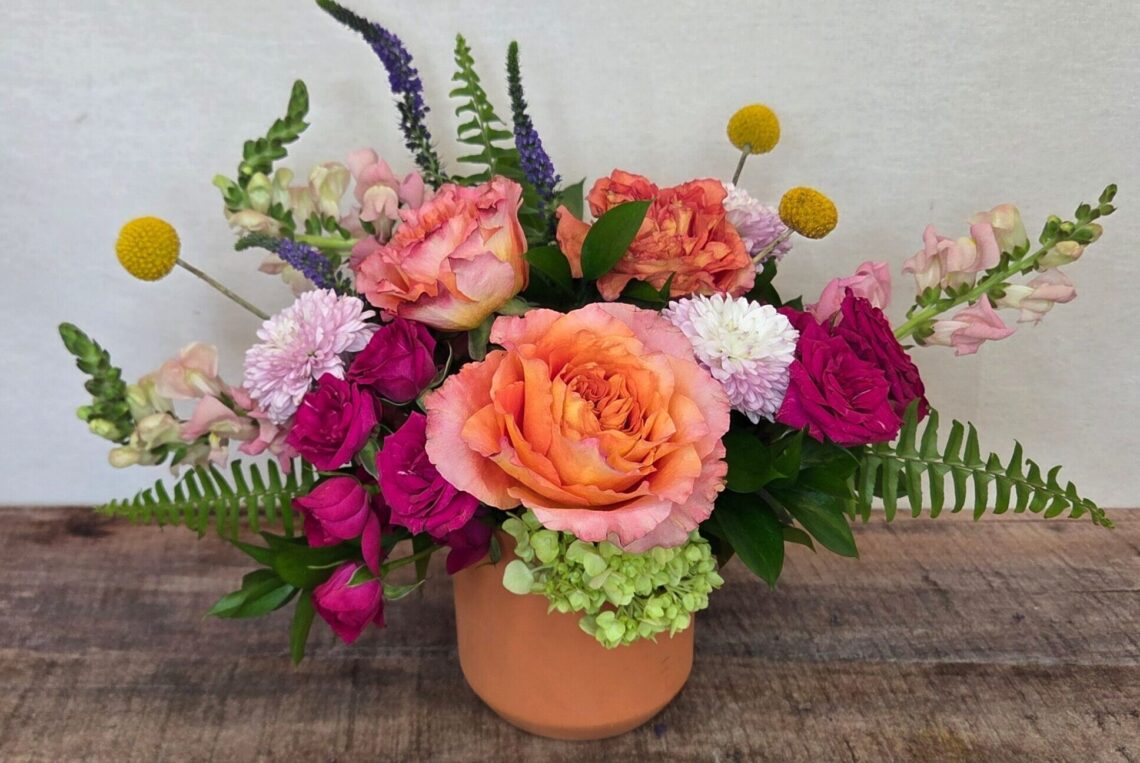 Apricot Smoothie is a cheerful arrangement with orangey apricot roses, hot pink spray roses, pastel snap dragons, and yellow Billy balls in a pastel container