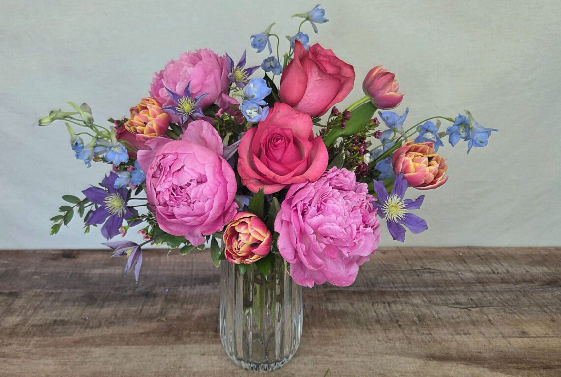 Blue Sunset deulxe ize features hot pink roses, bright pink peonies, with blue delphinium, tulips, and clematis in a glass vase
