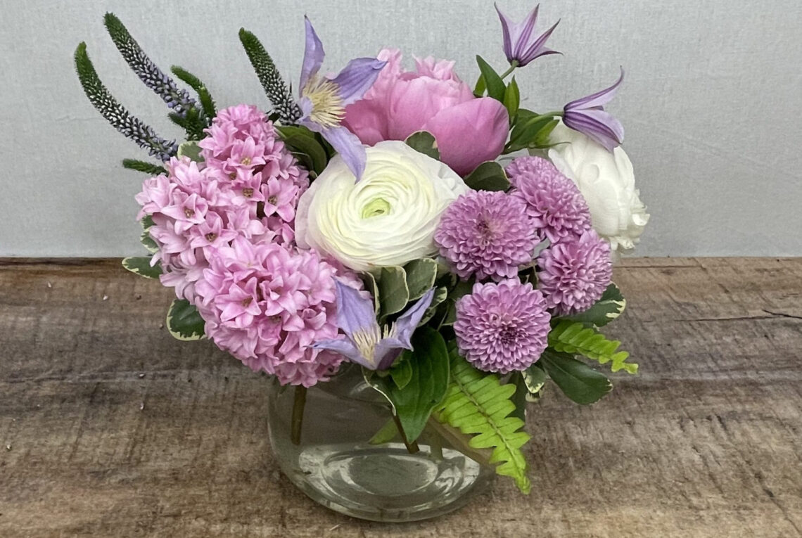 Cottage Darling is a charming mix of white and light purple blooms featuring ranunculus, hyacinth, clematis and veronica in a glass container