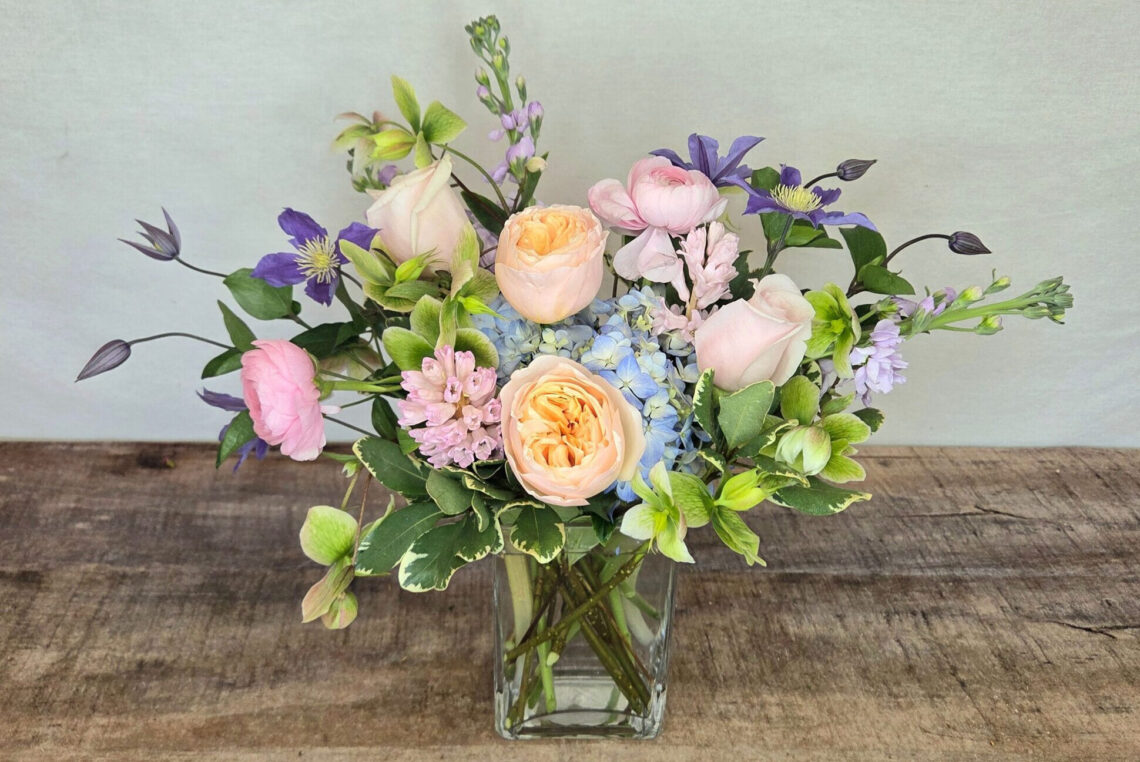 Dancin' Queen is a pastel mix of blooms featuring peach roses, blue hydrangea, and purple clematis with greenery in a glass vase