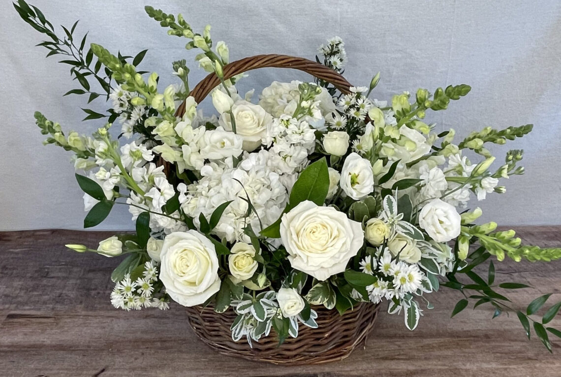 A Full Life sympathy arrangement in a basket in white monochromatic featuring roses and snapdragons