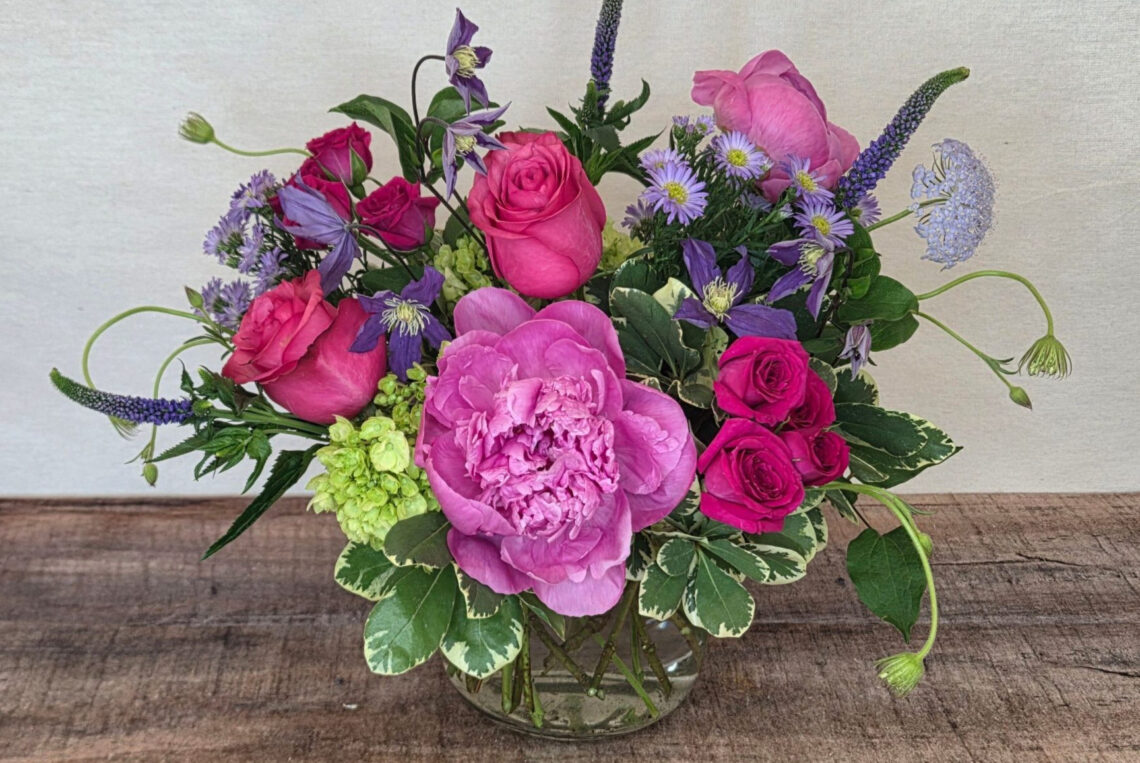 Gem Precious is a bright mix of hot pink peonies and roses with lavender and mint green accents with greenery in a glass container