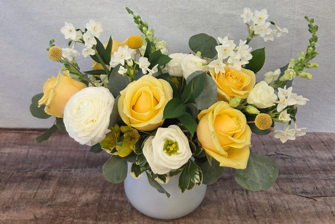 Hello, Buttercup features white and yellow blooms including roses and snapdragons in a white vase