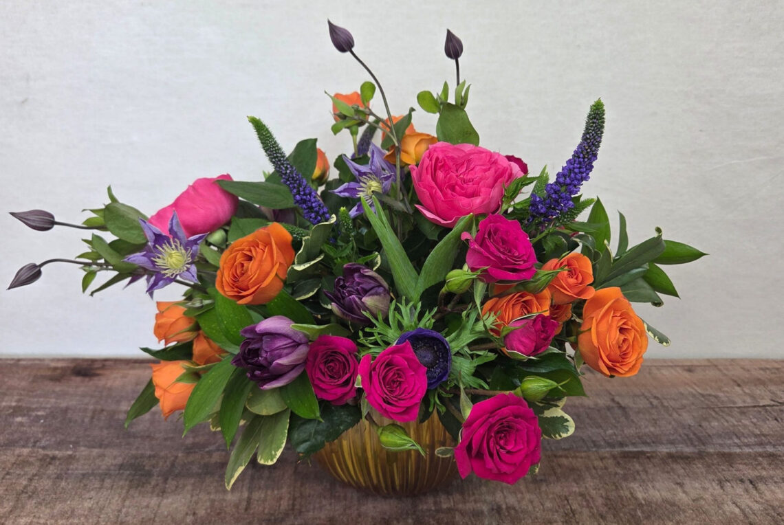 Joie De Vivre is a bright explosion of orange, hot pink, and purple blooms including roses, tulips, and veronica with greenery in a metallic bowl
