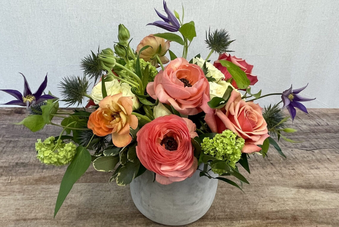 Ladybug is a mix of coral and peach ranunculus and roses with green viburnum and purple clematis and thistle in a rustic container