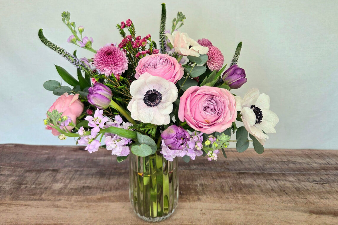 Lavender Waltz blends lavender, pink, and white blooms including ranunculus and anemones and tulips with greenery in a glass vase