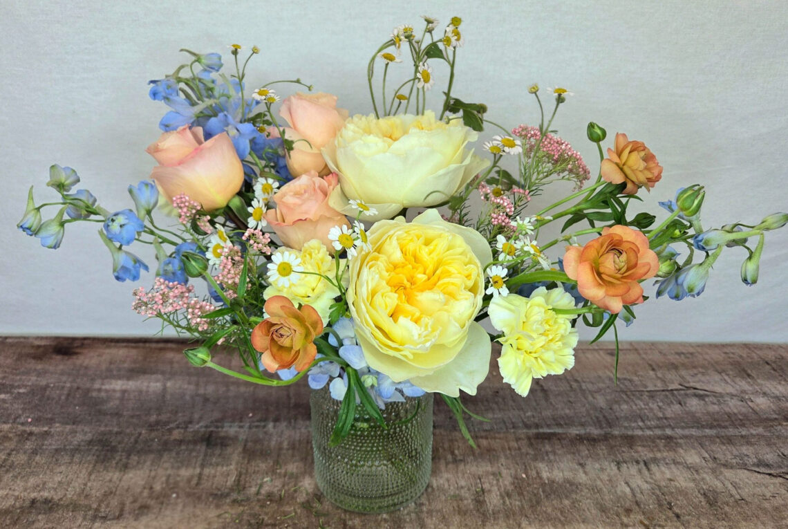 Ooo La Lemon is a pastel arrangement featuring lemon roses with peach accents and blue delphinium in a glass vase