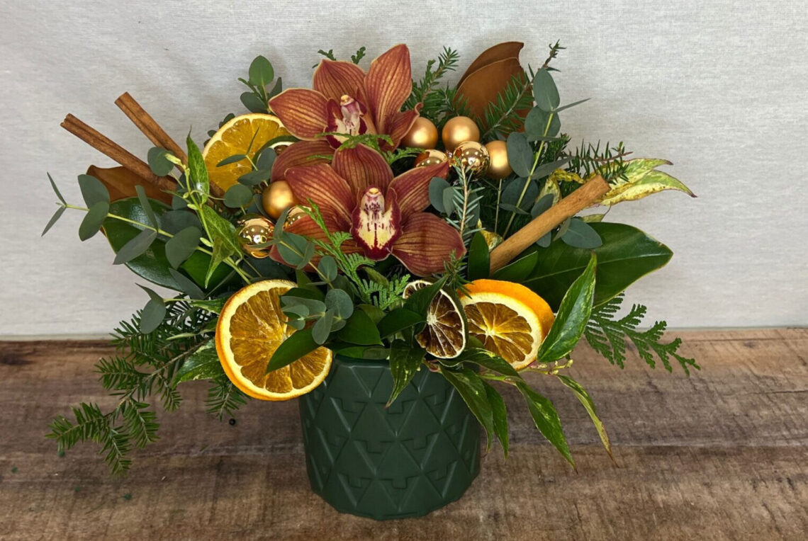 Orange You Merry is Charlie Brown orchids with orange slices, magnolia leaves, and evergreens in a deep green container