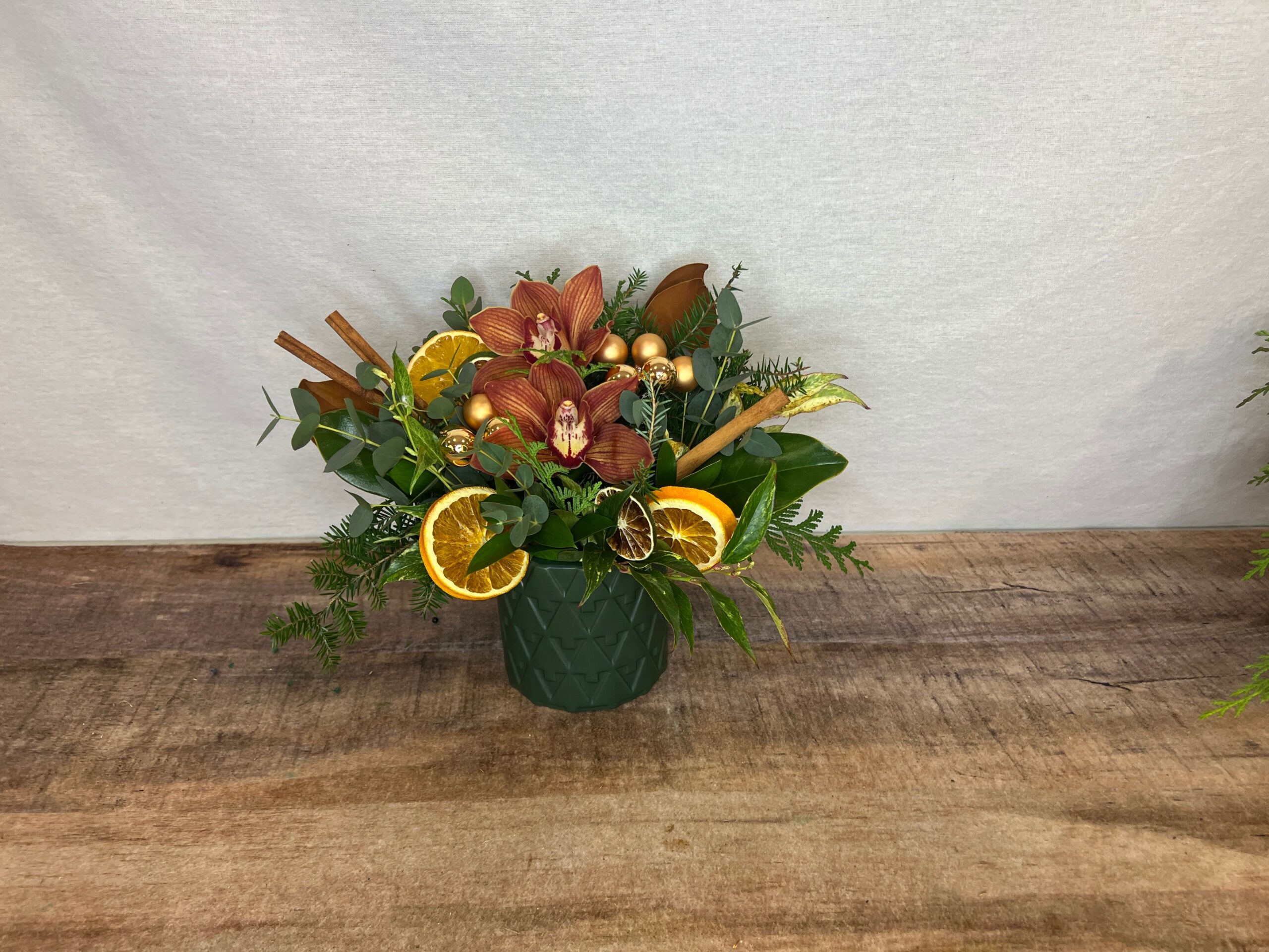 Orange You Merry is Charlie Brown orchids with orange slices, magnolia leaves, and evergreens in a deep green container