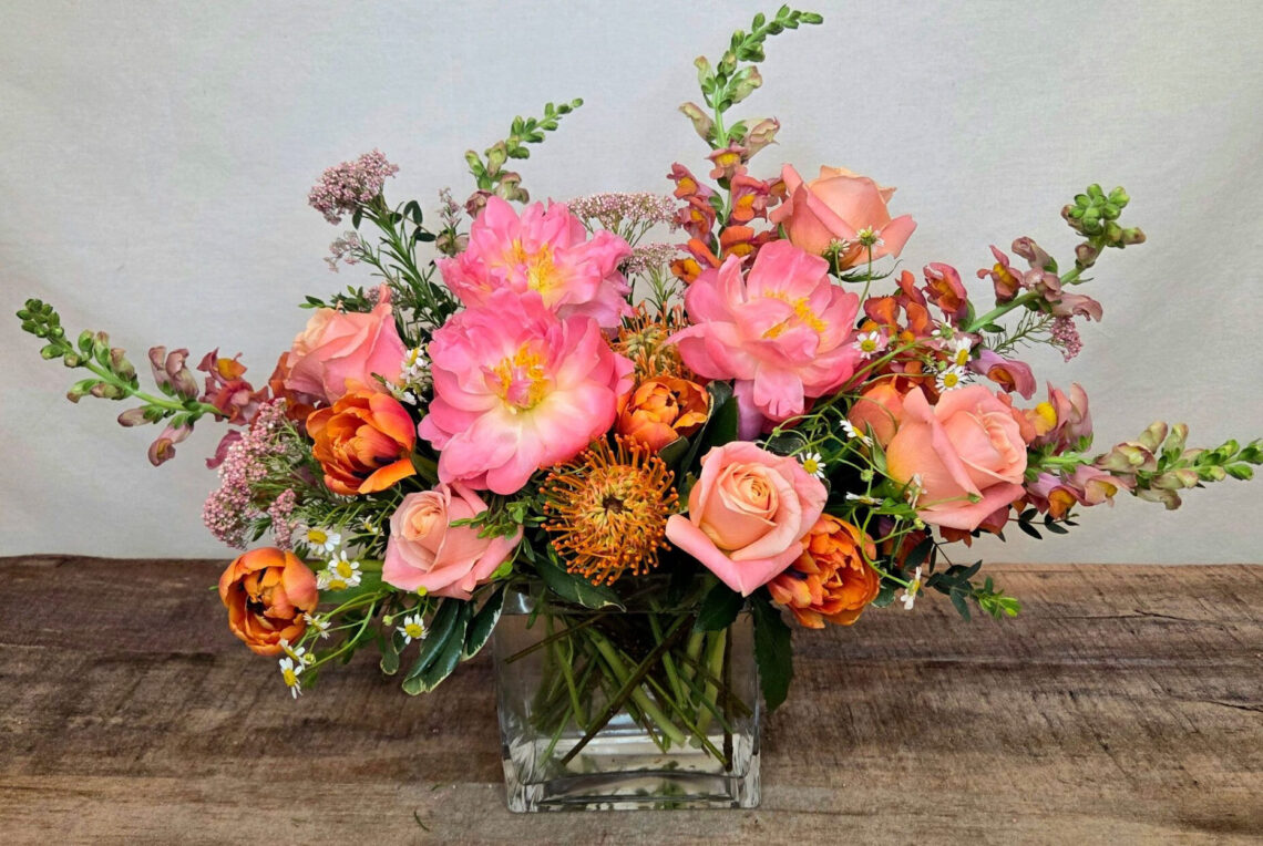 Peach Halo features Coral Charm peonies with roses, snap dragons, tulips, and an orange protea in a glass vase