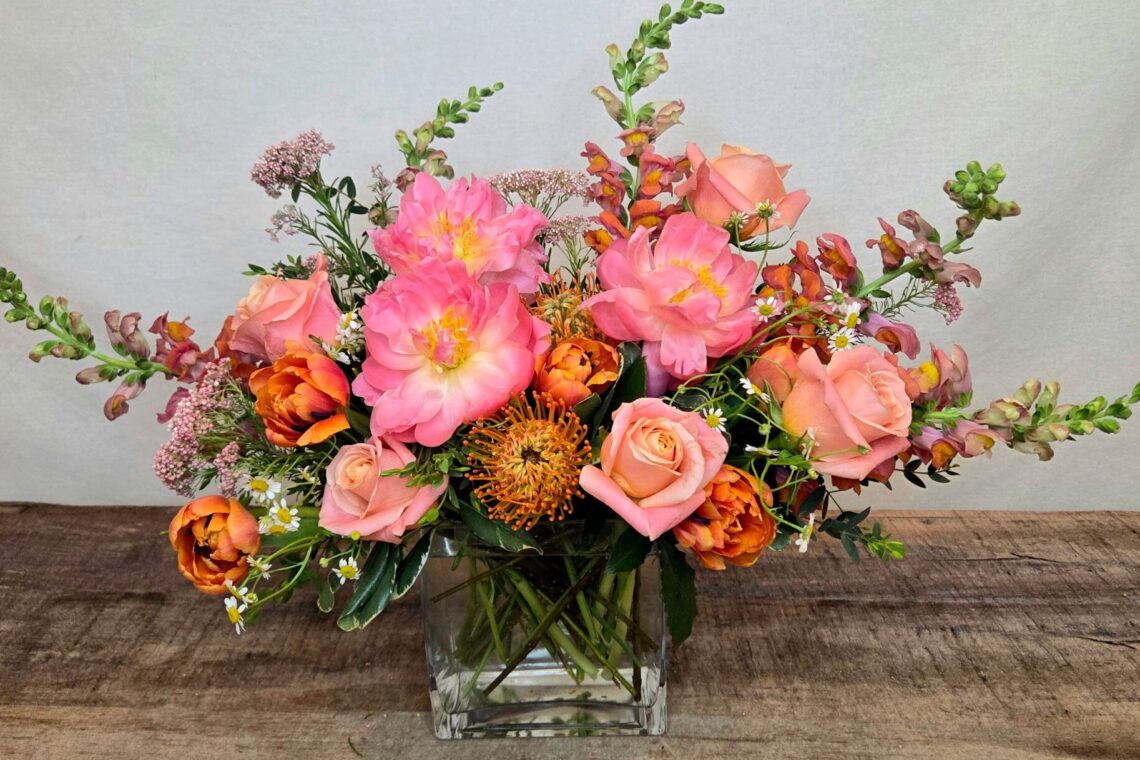 Peach Halo features Coral Charm peonies with roses, snap dragons, tulips, and an orange protea in a glass vase