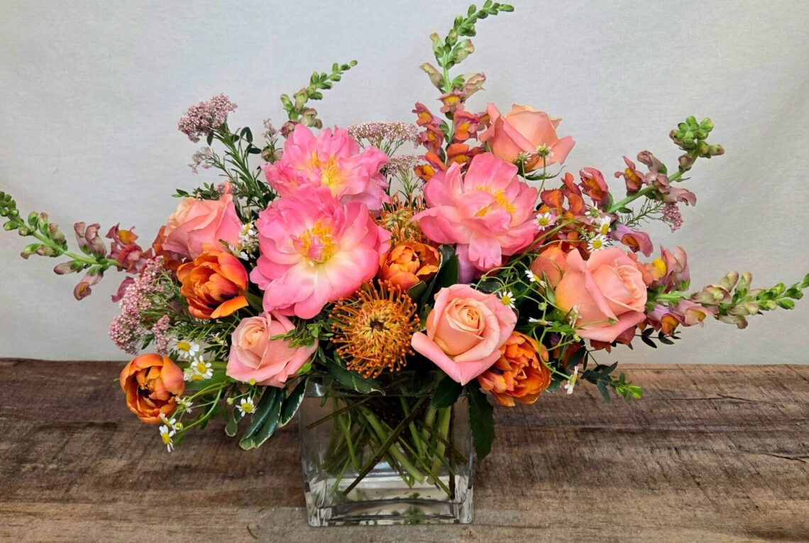 Peach Halo features Coral Charm peonies with roses, snap dragons, tulips, and an orange protea in a glass vase