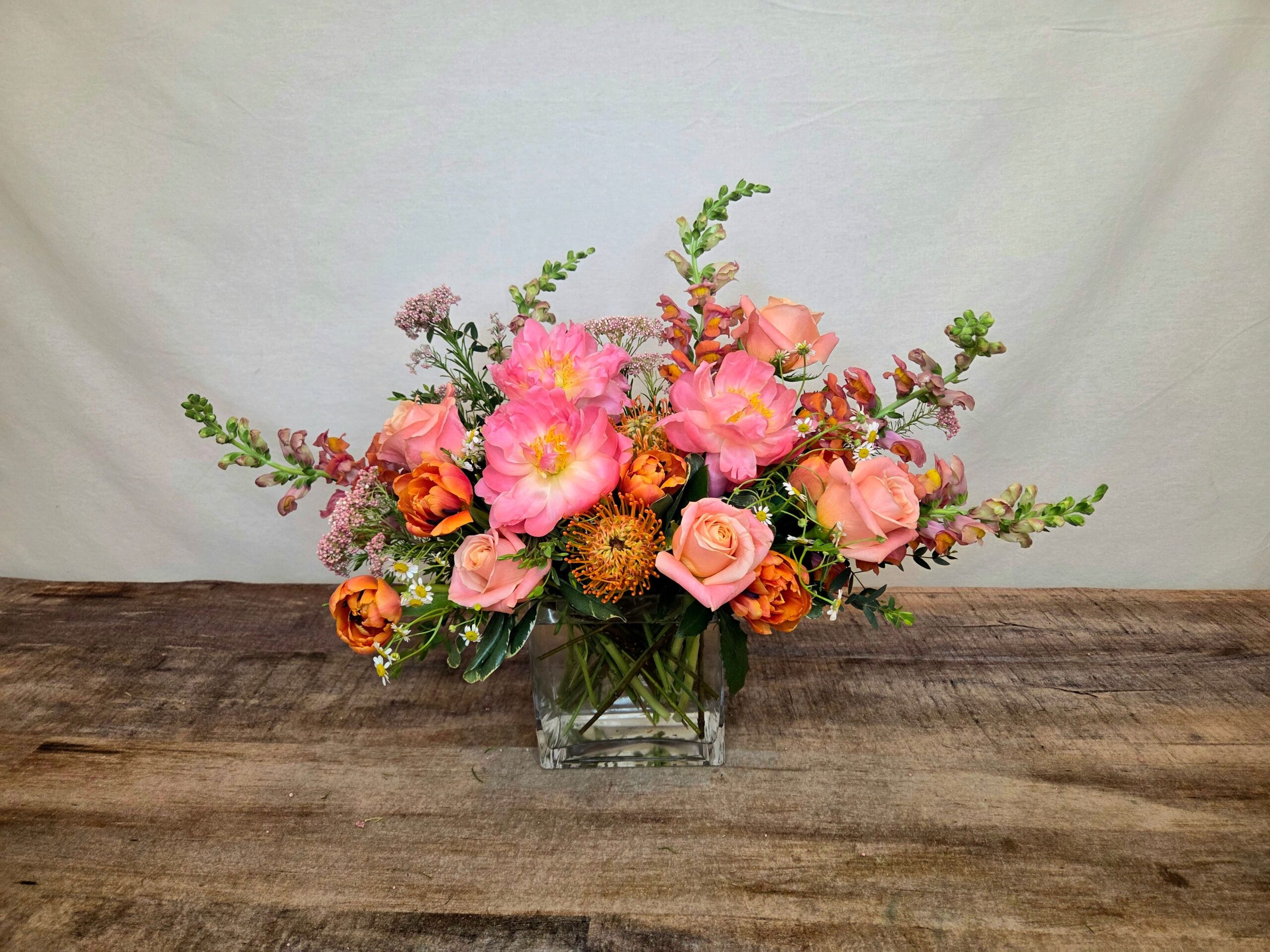 Peach Halo features Coral Charm peonies with roses, snap dragons, tulips, and an orange protea in a glass vase